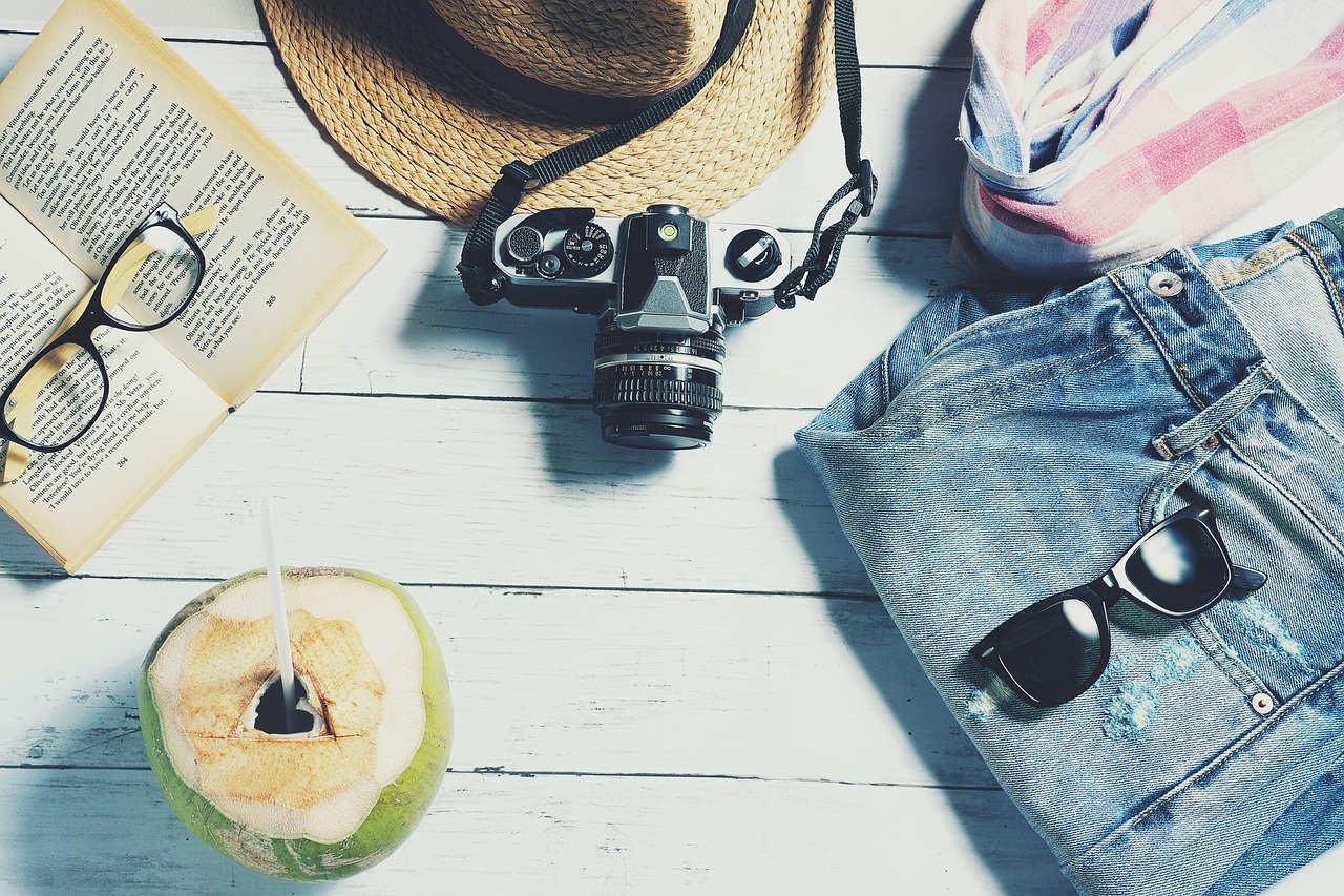 Jeans, sunglasses, coconut with straw, and DSLR camera laid out on wooden table